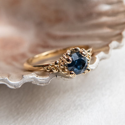 A gold ring with a textured band features a round brilliant cut deep blue sapphire, set in ornate gold prongs. 