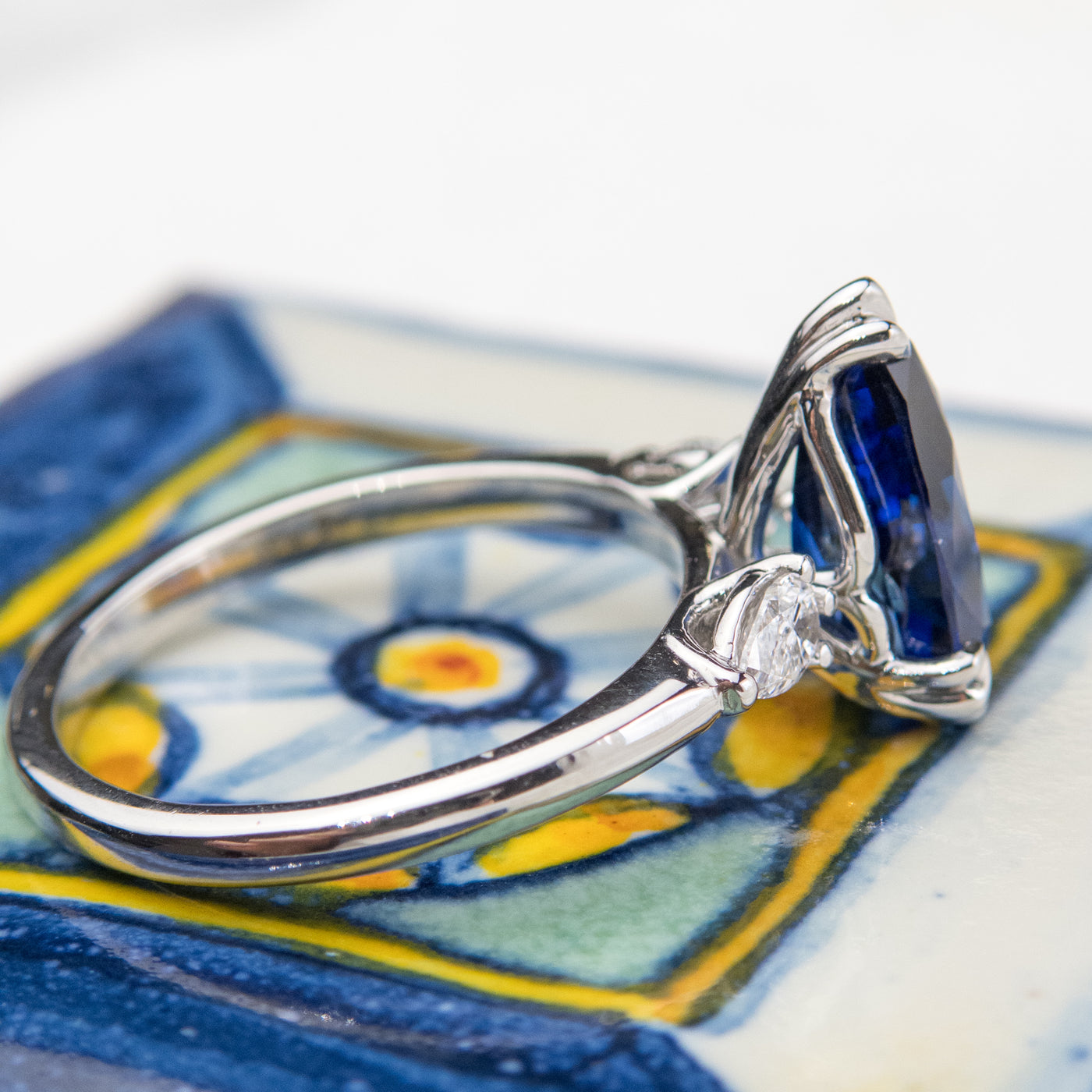 A side view of a pear-shaped blue sapphire engagement ring with white pear shaped diamond side stones, set in a platinum band, resting on a colourful blue and yellow surface.