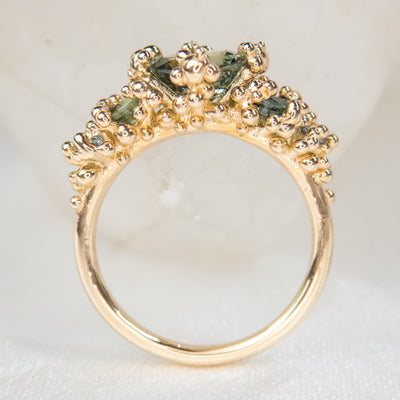 Side profile view of a gold ring featuring five green sapphires, surrounded by textured gold granule accents, set against a soft white backdrop.