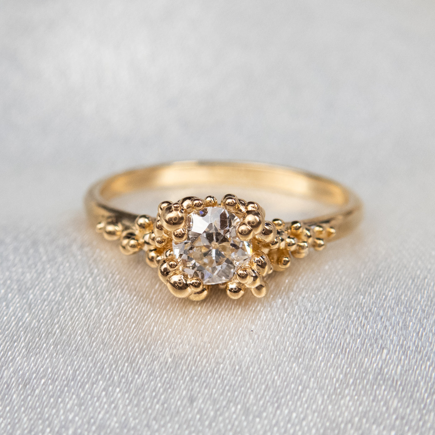 A gold ring featuring a central sparkling round old European cut diamond, surrounded by gold granules on a soft white fabric background. 