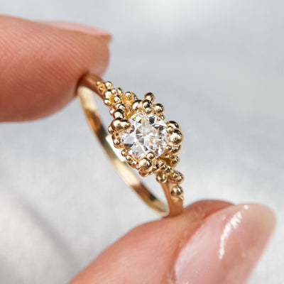 A gold ring featuring a central sparkling round old European cut diamond, surrounded by gold granules held between two fingers.