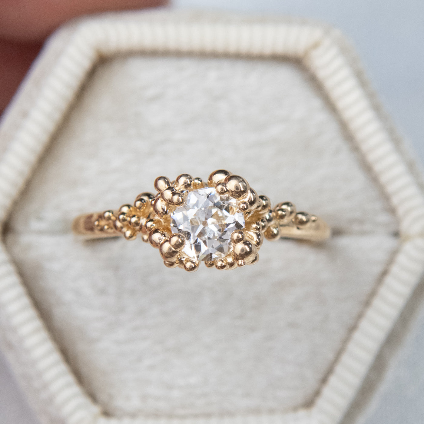 A gold ring featuring a central sparkling round old European cut diamond, surrounded by gold granules on a soft white fabric ring box.