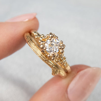 A gold ring featuring a central sparkling round old European cut diamond, surrounded by gold granules, stacked with a contoured organically textured wedding band.