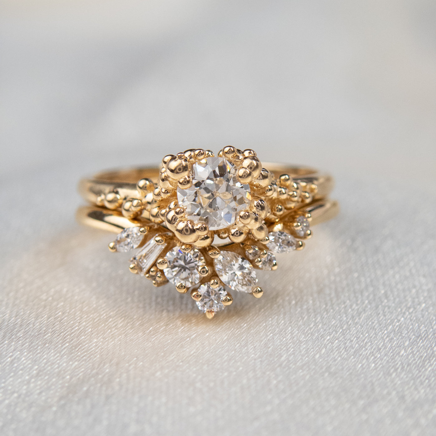 A gold ring featuring a central sparkling round old European cut diamond, surrounded by gold granules, stacked with a contoured diamond wedding band.