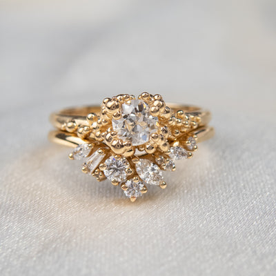 A gold ring featuring a central sparkling round old European cut diamond, surrounded by gold granules, stacked with a contoured diamond wedding band.