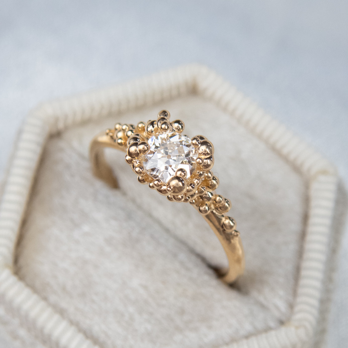 A gold ring featuring a central sparkling round old European cut diamond, surrounded by gold granules on a soft white fabric ring box.