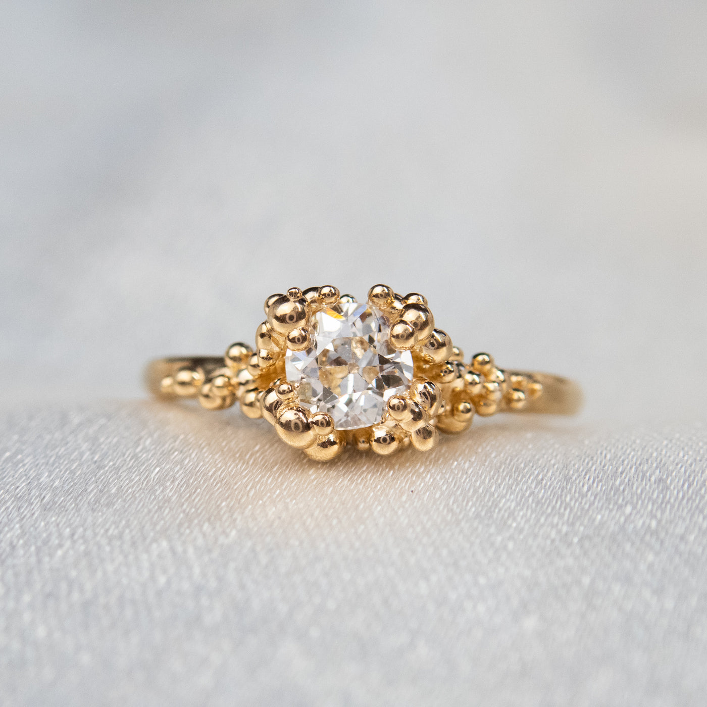 A gold ring featuring a central sparkling round old European cut diamond, surrounded by gold granules on a soft white fabric background. 