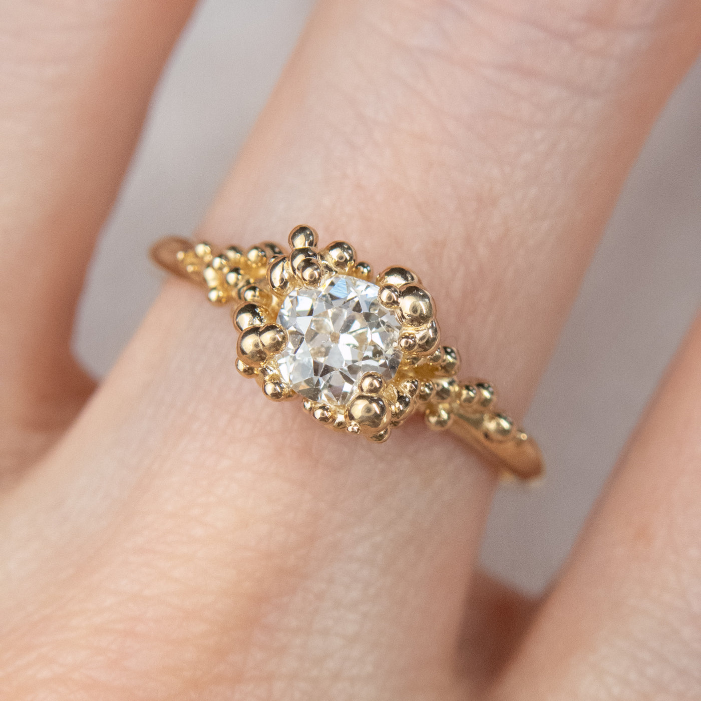 Top view of a gold ring featuring a central sparkling round old European cut diamond, surrounded by gold granules worn on a finger.