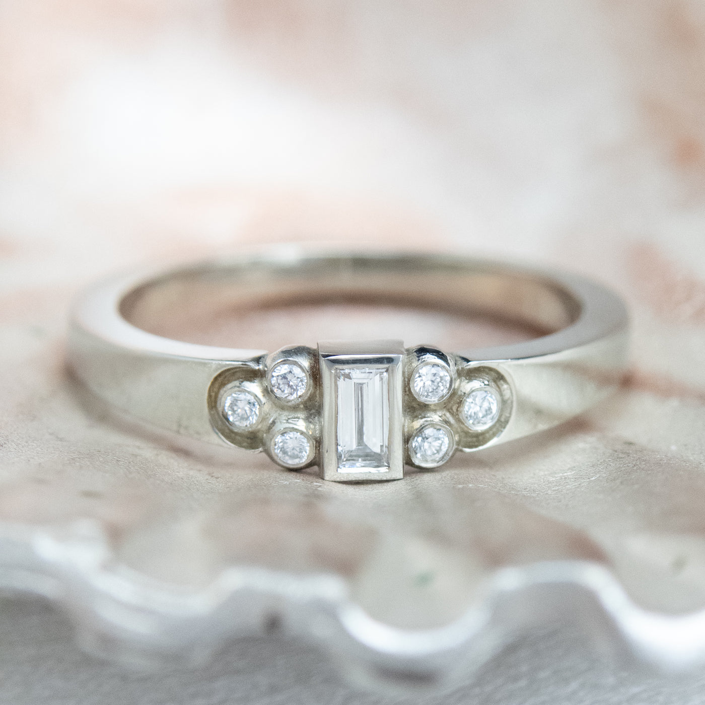 Elegant silver ring with a central rectangular-cut diamond flanked by six small round diamonds, set on a textured metallic surface. 