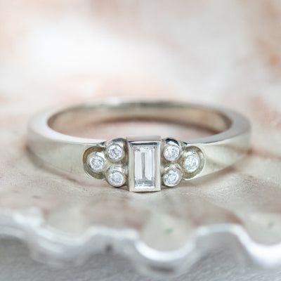 Elegant silver ring with a central rectangular-cut diamond flanked by six small round diamonds, set on a textured metallic surface. 