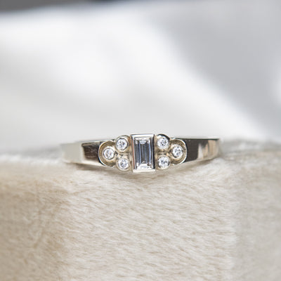 Elegant white gold ring with a central rectangular-cut diamond flanked by six small round diamonds, set on a textured white surface. 