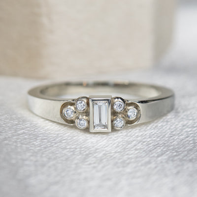 Elegant white gold ring with a central rectangular-cut diamond flanked by six small round diamonds, set on a textured white surface. 