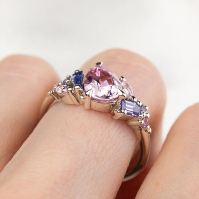 A white gold cluster ring featuring a large, pear-shaped pink sapphire at the center, surrounded by smaller, multi-shaped purple and lavender sapphires, worn on a finger.