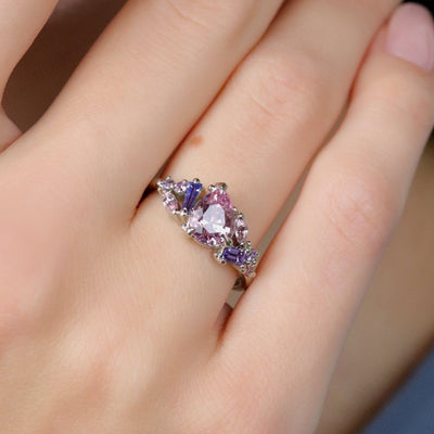 A white gold cluster ring featuring a large, pear-shaped pink sapphire at the center, surrounded by smaller, multi-shaped purple and lavender sapphires, worn on a hand.