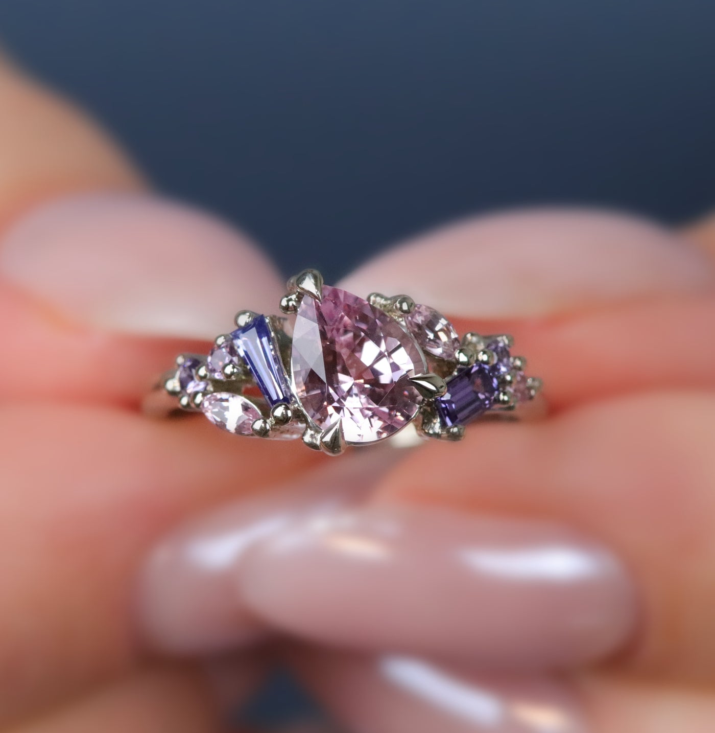 A white gold cluster ring featuring a large, pear-shaped pink sapphire at the center, surrounded by smaller, multi-shaped purple and lavender sapphires, being held by hands. 
