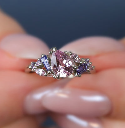 A white gold cluster ring featuring a large, pear-shaped pink sapphire at the center, surrounded by smaller, multi-shaped purple and lavender sapphires, being held by hands. 