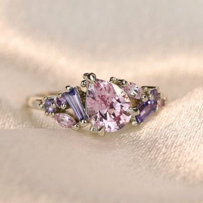 A white gold cluster ring featuring a large, pear-shaped pink sapphire at the center, surrounded by smaller, multi-shaped purple and lavender sapphires, on a soft fabric background. 