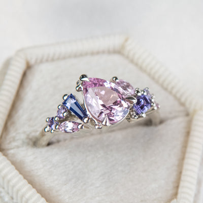 A white gold cluster ring featuring a large, pear-shaped pink sapphire at the center, surrounded by smaller, multi-shaped purple and lavender sapphires, in a white hexagonal shaped velvet ring box.