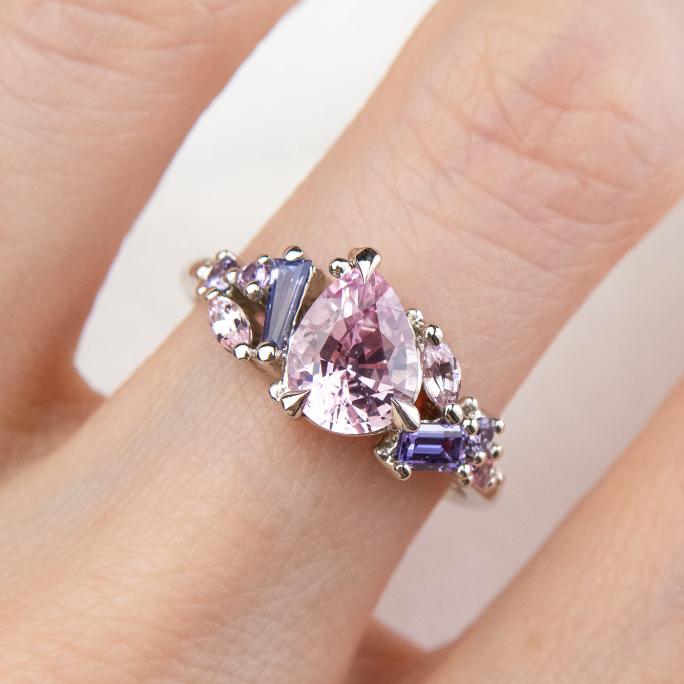 A white gold cluster ring featuring a large, pear-shaped pink sapphire at the center, surrounded by smaller, multi-shaped purple and lavender sapphires, worn on a finger.