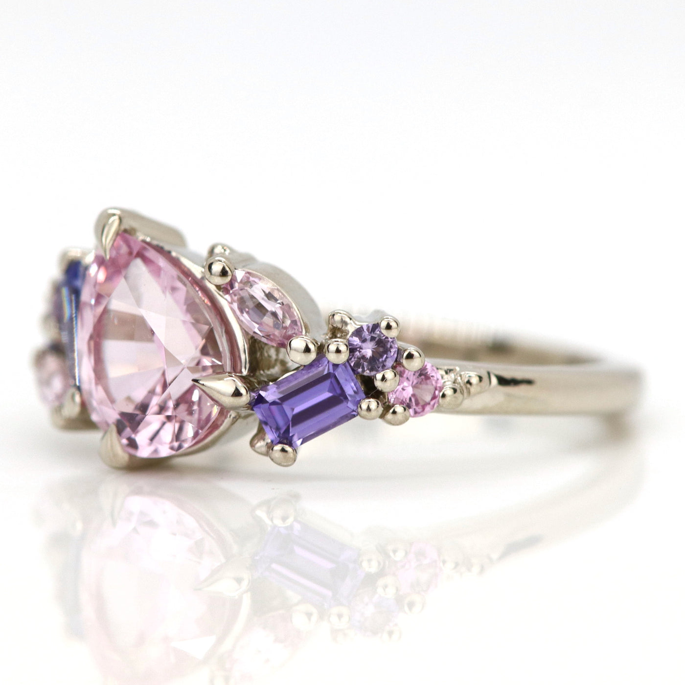 A white gold cluster ring featuring a large, pear-shaped pink sapphire at the center, surrounded by smaller, multi-shaped purple and lavender sapphires, on a reflective white background. 