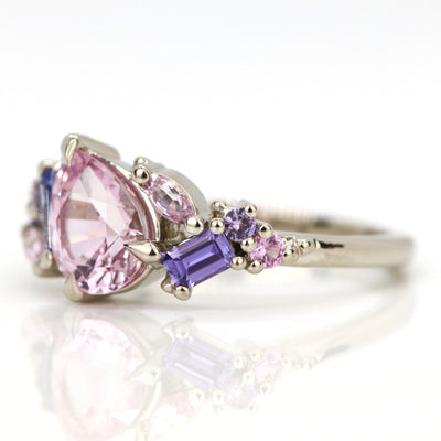 A white gold cluster ring featuring a large, pear-shaped pink sapphire at the center, surrounded by smaller, multi-shaped purple and lavender sapphires, on a reflective white background. 