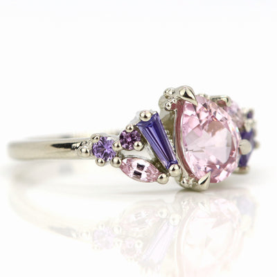 A white gold cluster ring featuring a large, pear-shaped pink sapphire at the center, surrounded by smaller, multi-shaped purple and lavender sapphires, on a reflective white background. 