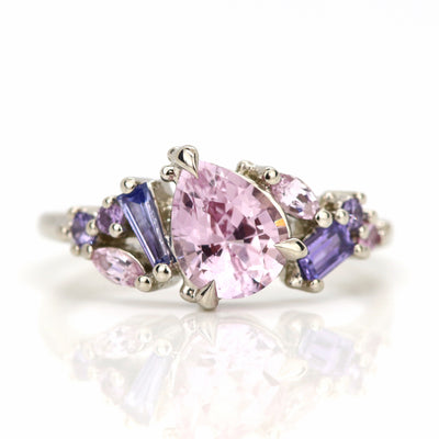 A white gold cluster ring featuring a large, pear-shaped pink sapphire at the center, surrounded by smaller, multi-shaped purple and lavender sapphires, on a reflective white background. 