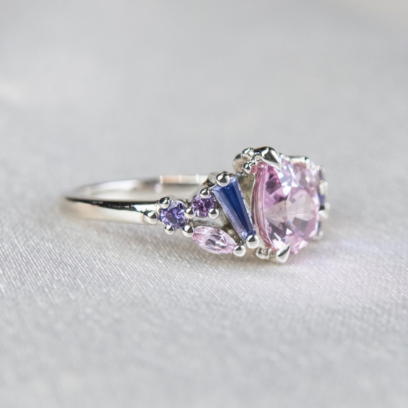 A white gold cluster ring featuring a large, pear-shaped pink sapphire at the center, surrounded by smaller, multi-shaped purple and lavender sapphires, set on a soft white fabric.