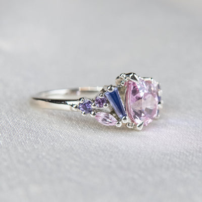A white gold cluster ring featuring a large, pear-shaped pink sapphire at the center, surrounded by smaller, multi-shaped purple and lavender sapphires, set on a soft white fabric.