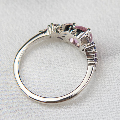 A white gold cluster ring featuring a large, pear-shaped pink sapphire at the center, surrounded by smaller, multi-shaped purple and lavender sapphires, set on a soft white fabric.