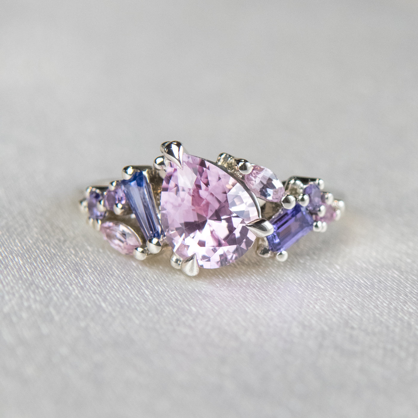 A white gold cluster ring featuring a large, pear-shaped pink sapphire at the center, surrounded by smaller, multi-shaped purple and lavender sapphires, set on a soft white fabric.