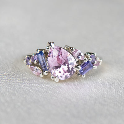 A white gold cluster ring featuring a large, pear-shaped pink sapphire at the center, surrounded by smaller, multi-shaped purple and lavender sapphires, set on a soft white fabric.