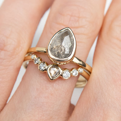A delicate gold ring with a central pear-shaped diamond, flanked by smaller round diamonds and worn with a bezel set pear engagement ring worn on a finger.