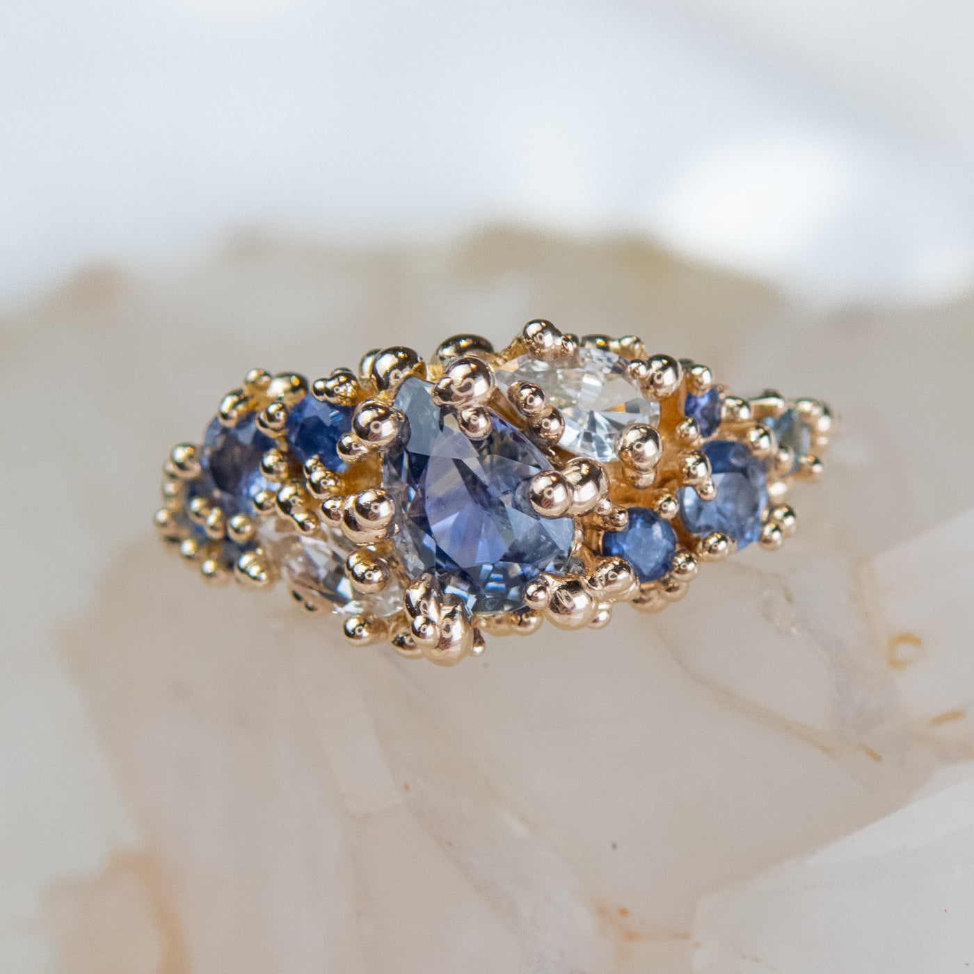 Intricately designed gold cluster ring featuring blue and colourless sapphires, set atop a crystalline backdrop.