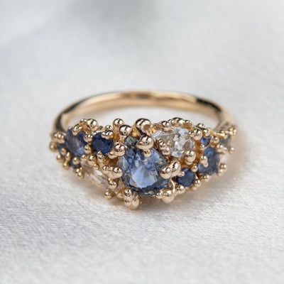 Intricately designed gold cluster ring featuring blue and colourless sapphires, set atop a white fabric backdrop.