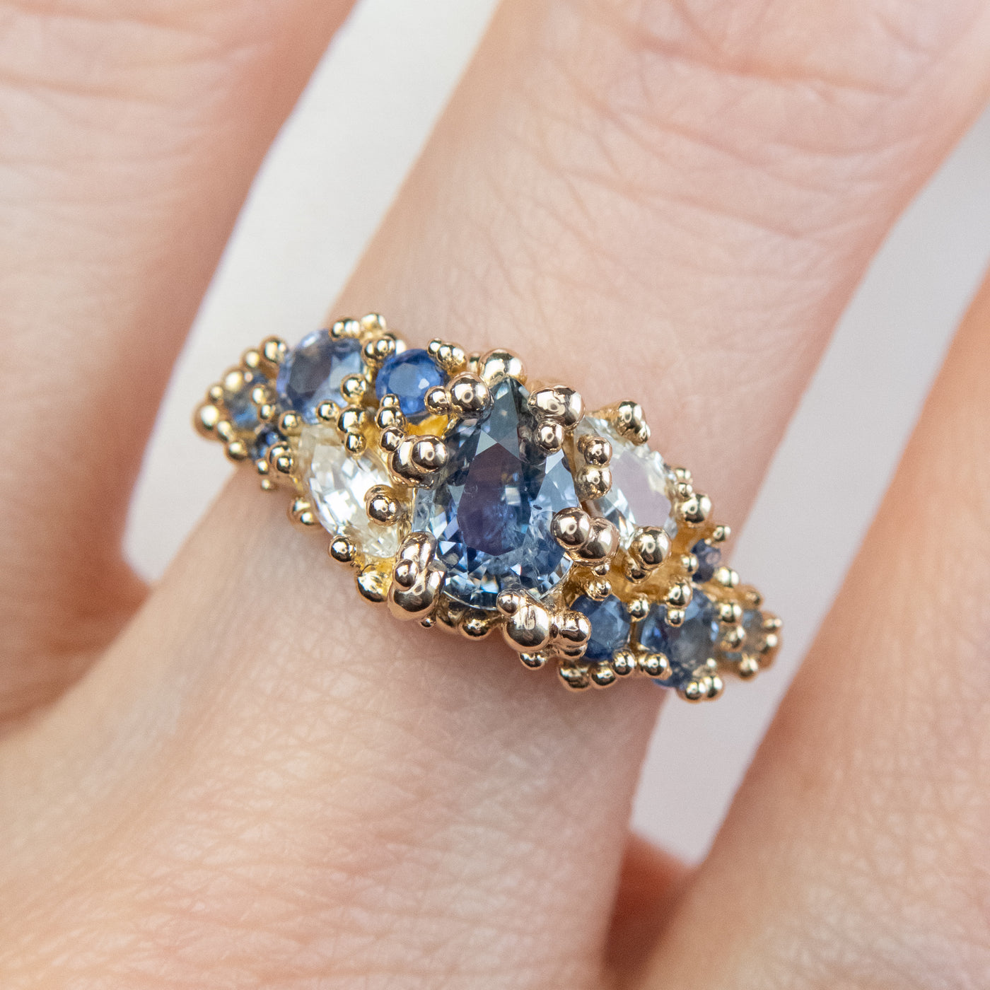 Intricately designed gold cluster ring featuring blue and colourless sapphires, worn on a finger. 