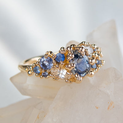 Intricately designed gold cluster ring featuring blue and colourless sapphires, set atop a crystalline backdrop.