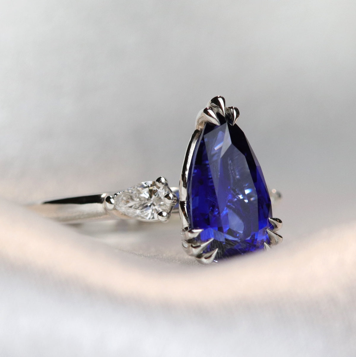 A pear-shaped blue sapphire ring with white pear shaped diamond side stones, set in a platinum band, resting on a soft, light grey fabric.