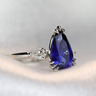 A pear-shaped blue sapphire ring with white pear shaped diamond side stones, set in a platinum band, resting on a soft, light grey fabric.