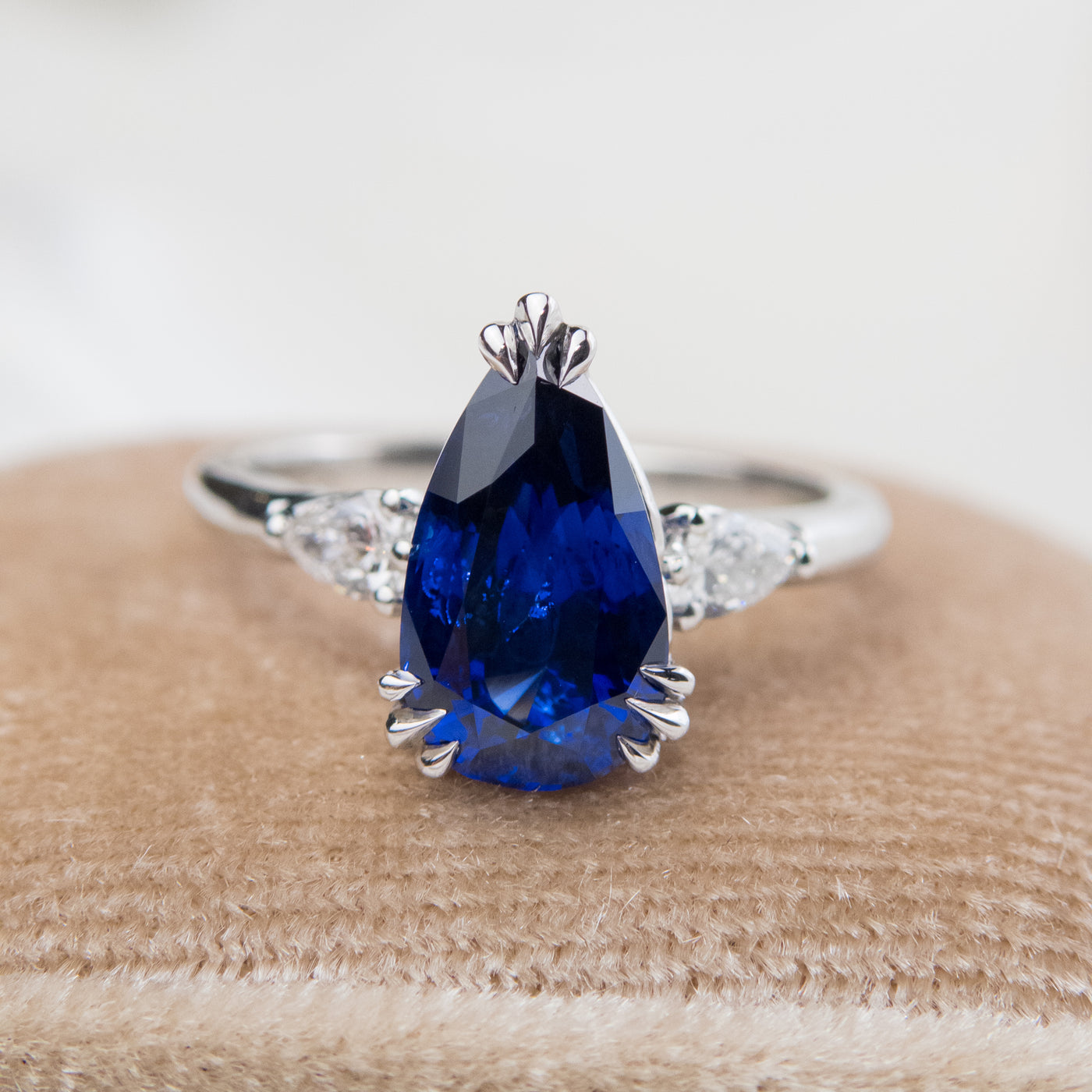 A pear-shaped blue sapphire ring with white pear shaped diamond side stones, set in a platinum band, resting on a soft, light fabric.