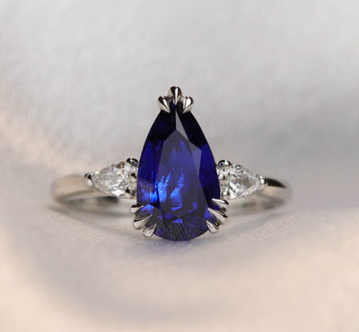 A pear-shaped blue sapphire ring with white pear shaped diamond side stones, set in a platinum band, resting on a soft, light grey fabric.