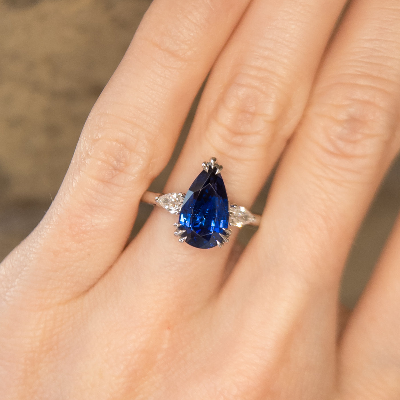 A pear-shaped blue sapphire ring with white pear shaped diamond side stones, set in a platinum band, worn on a hand. 