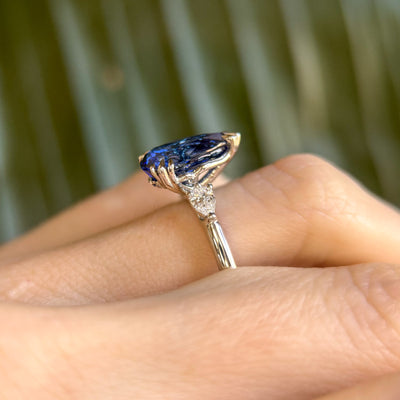 A pear-shaped blue sapphire ring with white pear shaped diamond side stones, set in a platinum band, worn on a hand. 