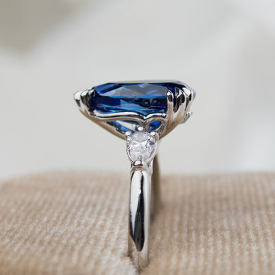 A side view of a pear-shaped blue sapphire engagement ring with white pear shaped diamond side stones, set in a platinum band, sitting in a soft velvet ring box.