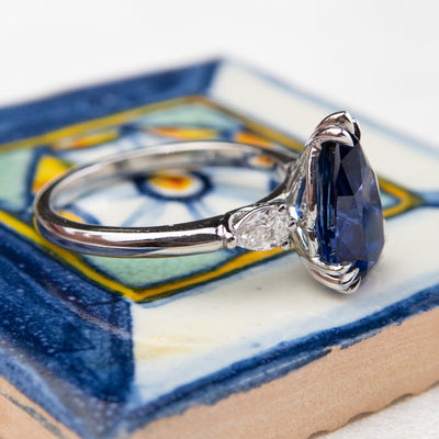 A side view of a pear-shaped blue sapphire engagement ring with white pear shaped diamond side stones, set in a platinum band, resting on a colourful blue and yellow surface.