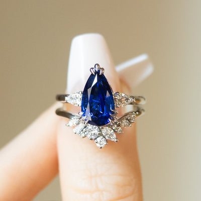 A pear-shaped blue sapphire ring with white pear shaped diamond side stones, set in a platinum band, paired with an intricate diamond band, held between two fingers.