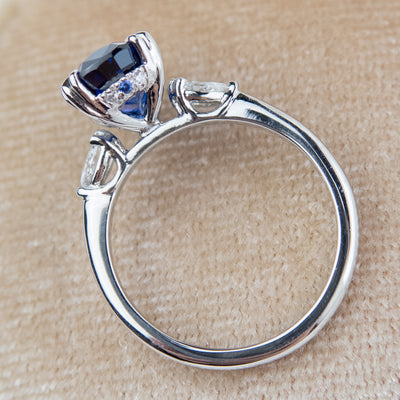 A side view of a pear-shaped blue sapphire engagement ring with white pear shaped diamond side stones, set in a platinum band, sitting on a soft velvet surface. 