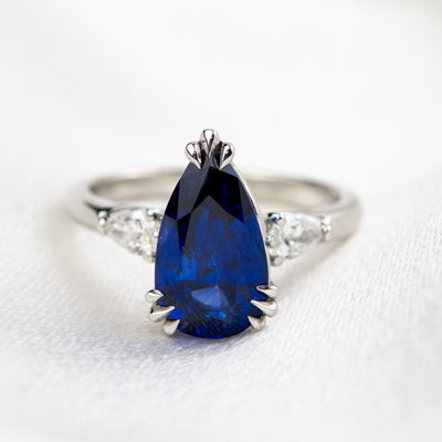 A pear-shaped blue sapphire ring with white pear shaped diamond side stones, set in a platinum band, resting on soft, light fabric.