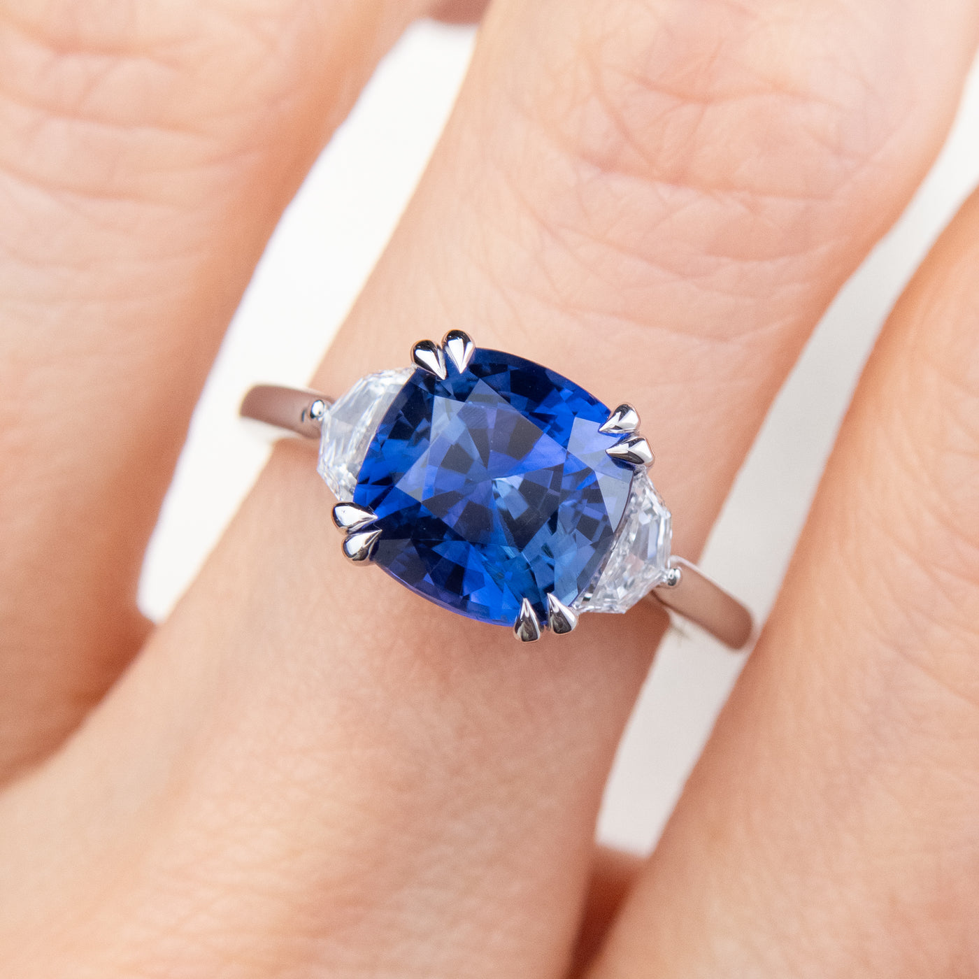 A large cushion shaped blue sapphire ring flanked by white cadillac cut diamonds set in a classic platinum setting, worn on a finger. 