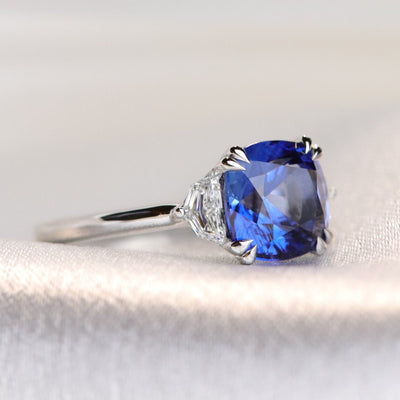A large cushion shaped blue sapphire ring flanked by white cadillac cut diamonds set in a classic platinum setting against a light, softly blurred background. 